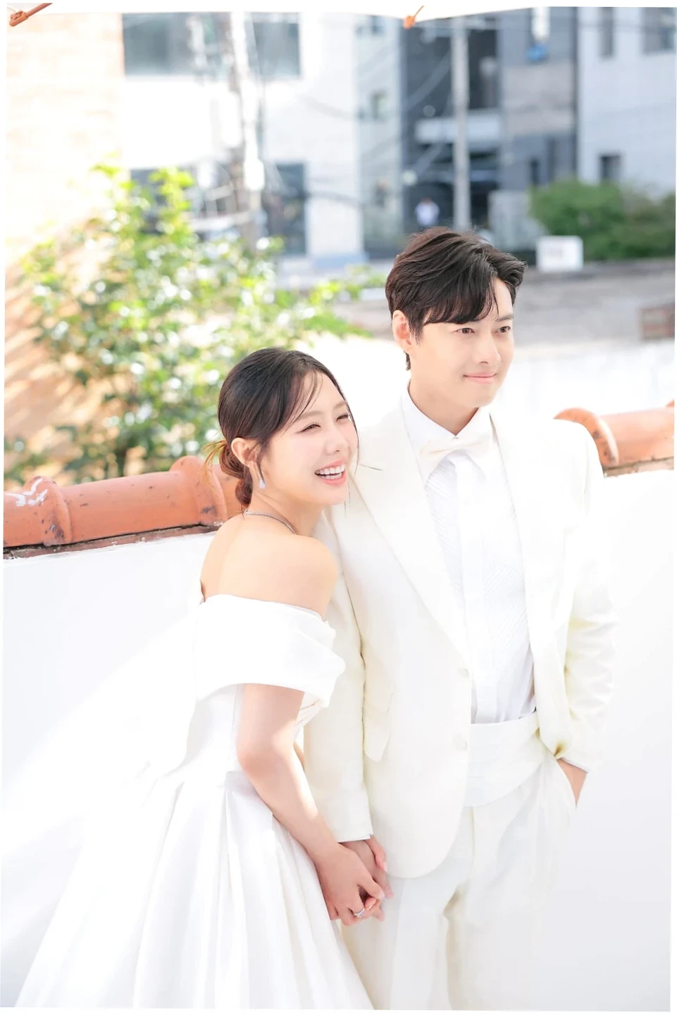 환하게 웃는 신부와 함께 팔짱을 끼고 손을 맞잡은 신랑의 모습. 하얀 드레스와 신랑예복을 입은 두 사람이 행복하게 서로를 바라보고 있다.