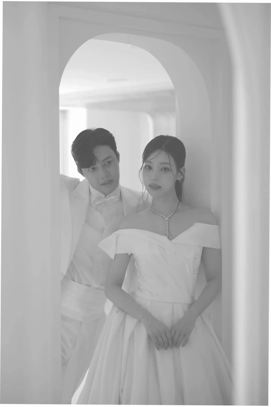 아치형 문 안에서 흰색 턱시도 차림의 신랑이 오프숄더 웨딩드레스와 목걸이를 한 신부 뒤에 서 있는 흑백 신랑예복 사진.