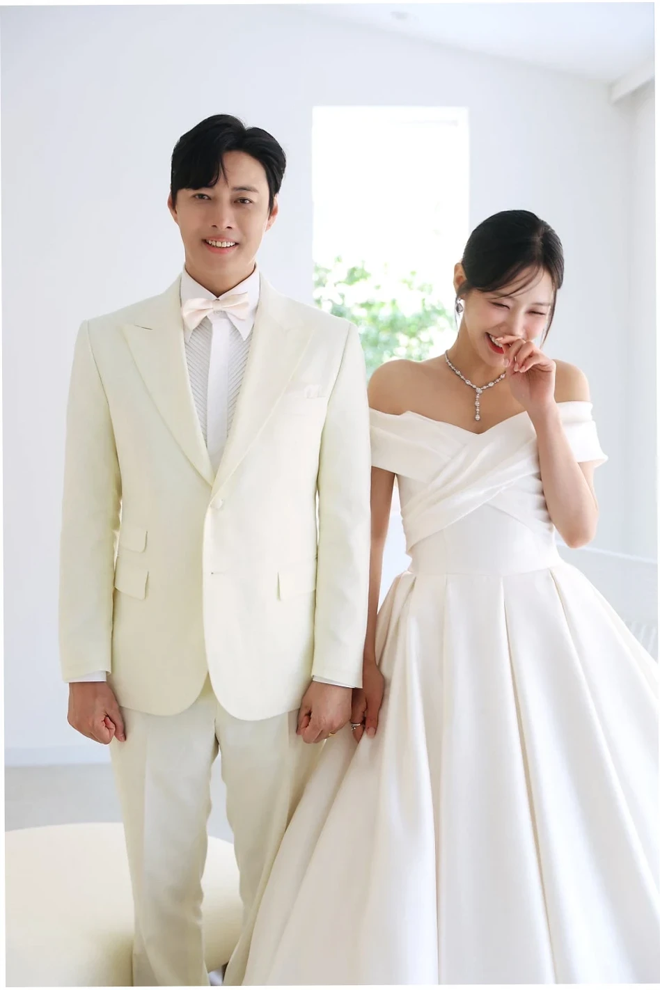밝은 아이보리 톤의 신랑예복을 입은 신랑과 순백의 웨딩드레스를 입은 신부가 손을 잡고 행복한 미소를 짓고 있다. 신부는 살짝 고개 숙여 웃고 있고, 신랑은 카메라를 보며 환하게 웃는 모습.